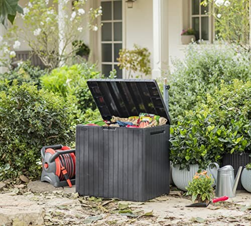 Open garden storage box with tools and hose near plants.